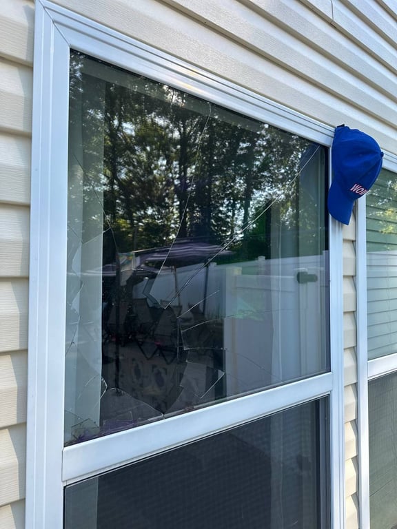 Broken residential window with cracked glass pane showing damage pattern, white vinyl frame against beige siding with tree reflections