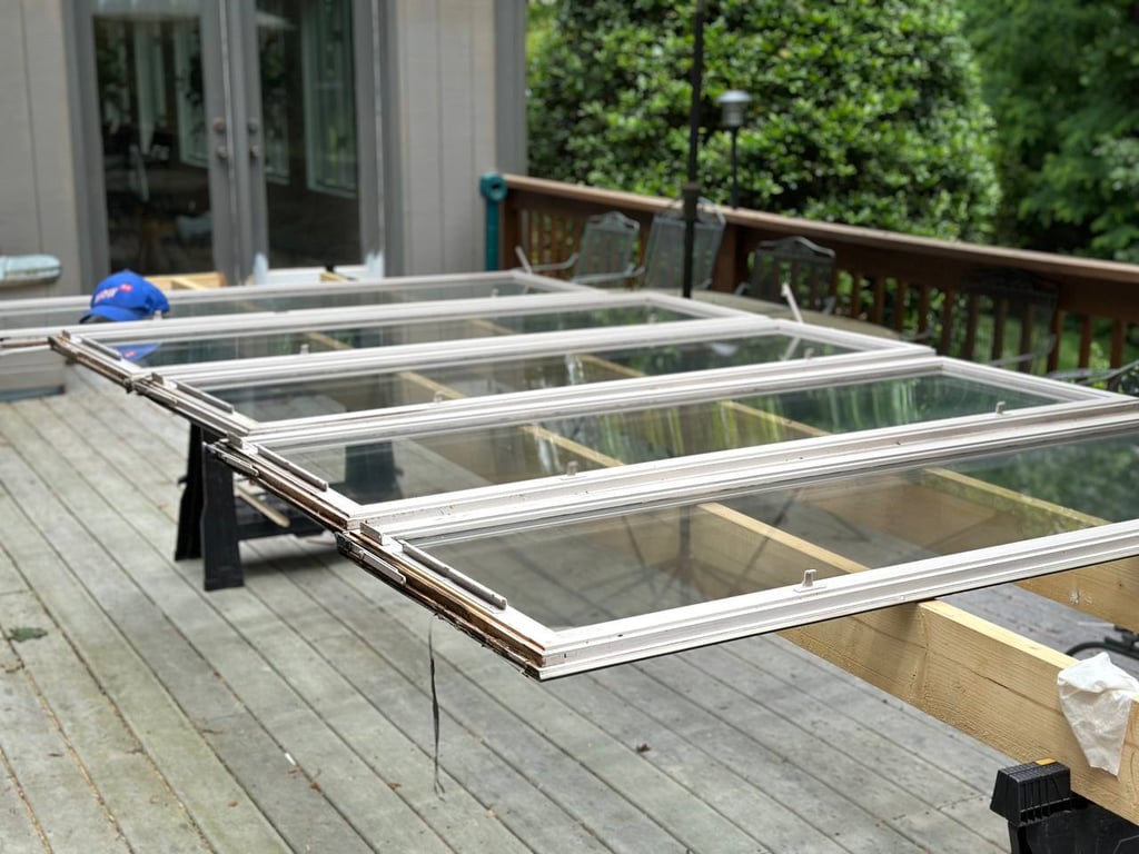 Window sash repair and restoration in progress showing multiple white-framed glass panels laid out on sawhorses on wooden deck