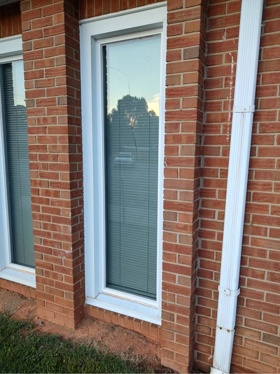Cracked full-length window with white trim in brick home exterior, showing residential window damage needing professional repair