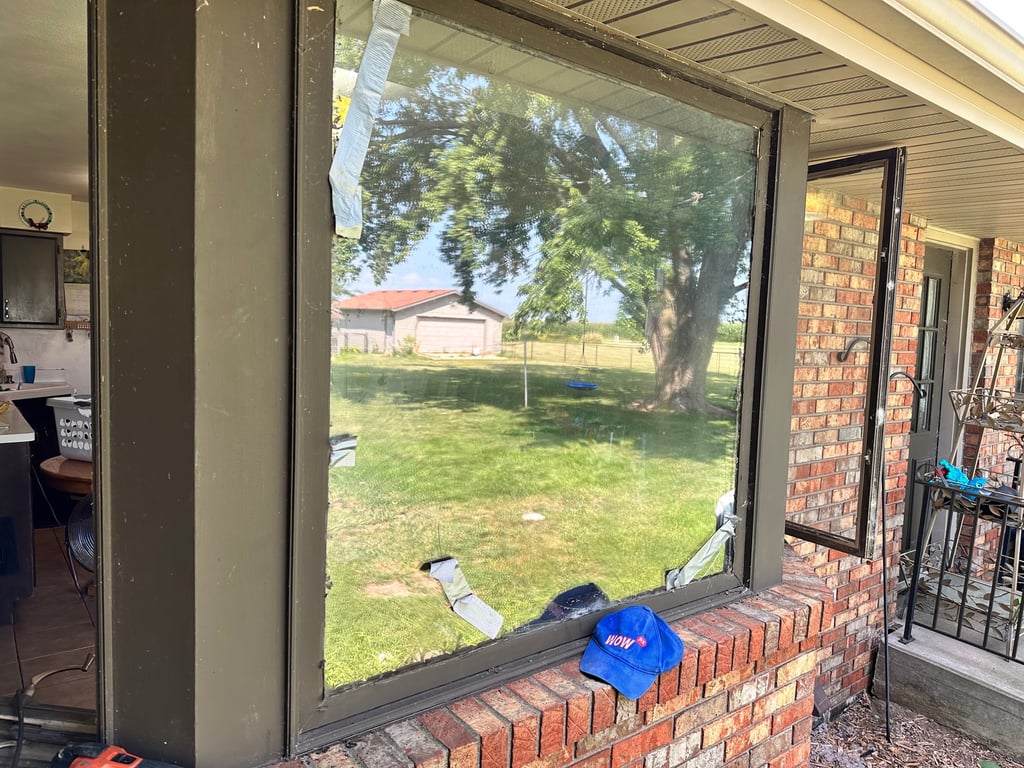 Broken residential window with WOW company hat showing lawn view, demonstrating need for emergency window repair in Greensboro